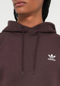 Artimas žmogaus apšvietimas, dėvinčio tamsiai purpurinę Adidas džemperį su baltu logotipu ant krūtinės ir iš šonų matomais pintais plaukais.