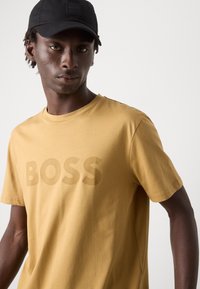 Mustard yellow T-shirt with a textured "BOSS" logo in a slightly darker shade. Short sleeves and crew neckline, made of soft fabric.