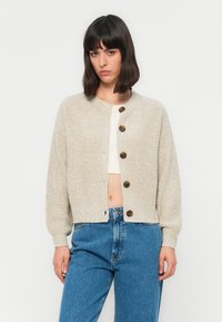 Kvinne med mørkt hår iført en beige strikket cardigan med knapper over en hvit kort topp og høyde midje blå jeans, som står mot en ensfarget bakgrunn.