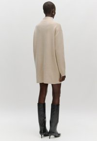 Beige trui-jurk met een hoge kraag, lange mouwen en een ontspannen pasvorm, gecombineerd met zwarte kniehoge laarzen met hak. Gladde textuur.