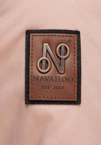 Hnedá kožená nálepka s vyrazeným logom "N" a nápisom "NAVAHOO EST 2014", obklopená čiernym prešívaním, na svetloružovom látkovom pozadí.