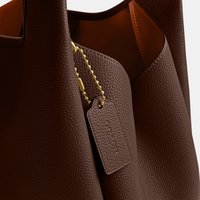 Borsa tote in pelle marrone texturizzata con interno liscio color cuoio chiaro e targhetta in pelle Coach appesa a una catena dorata con palline.