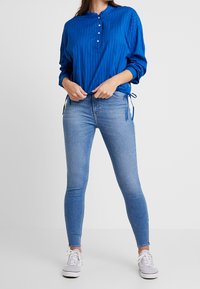 Camisa de botones a rayas azules, de corte relajado, combinada con jeans ajustados de un azul claro y zapatillas grises. La camisa presenta un cuello henley y se ata a los lados.