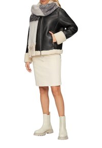 Veste en cuir noire avec des poignets en shearling crème, associée à une jupe crème claire et des bottines beiges. Une écharpe rayée grise et blanche complète le look.