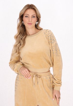 Kvinde med langt bølget blondt hår iført tan-farvet velour sweatshirt og bukser prydet med sølvnitter, poserer med hånden på hoften mod en hvid baggrund.