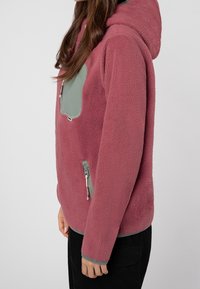 Kvinde iført en mauve fleecejakke med grønne lynlåslommer og hætte, der står sidelæns mod en ensfarvet baggrund.