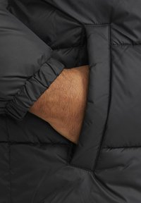 Jachetă puffer neagră din material cu un model matlasat. Prim-plan al unei manșete care arată un tiv elastic și o mână așezată într-un buzunar lateral.