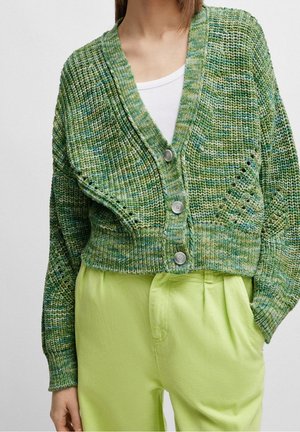 Kobieta ubrana w zapinany na guziki zielony dziergany kardigan na białym topie i jasnooliwkowych spodniach z wysokim stanem, z ręką w kieszeni, stojąca na jednolitym tle.