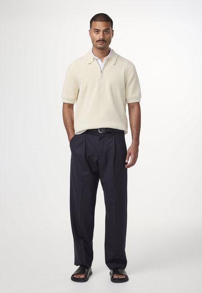 Creme kortærmet strik polo med lynlås fortil, parret med marineblå stribede bukser og sorte slidesandaler. Minimalistisk design og tekstureret stof.