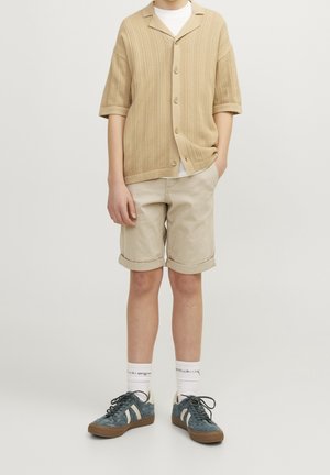 Person, der eine beigen Strick-Cardigan mit kurzen Ärmeln, ein weißes Unterhemd, beige gerollte Shorts, weiße Socken und blaue Sneakers trägt, steht vor einem einfarbigen Hintergrund.