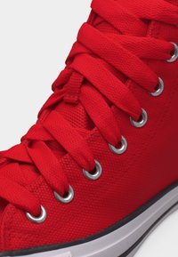 Κοντινή λήψη κόκκινου υφασμάτινου αθλητικού παπουτσιού με χοντρές κόκκινες κορδόνια διαπερασμένες από μεταλλικά κορδόνια και λευκή ελαστική σόλα.