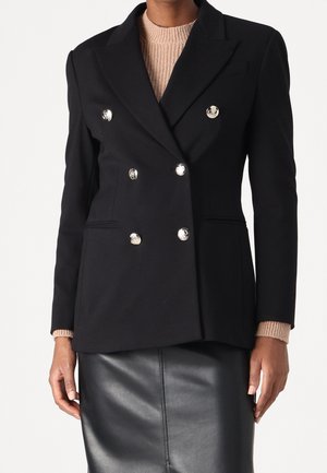 Personne portant un blazer croisé noir avec des boutons argentés brillants sur un pull en maille beige et une jupe en cuir noir, debout devant un fond blanc.