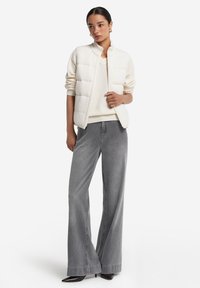 Leichte, gesteppte weiße Weste mit hohem Kragen, getragen über einem cremefarbenen Pullover und kombiniert mit hoch taillierten grauen weiten Jeans und schwarzen High Heels.