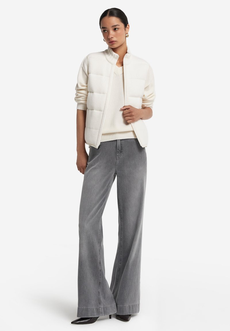 Leichte, gesteppte weiße Weste mit hohem Kragen, getragen über einem cremefarbenen Pullover und kombiniert mit hoch taillierten grauen weiten Jeans und schwarzen High Heels.