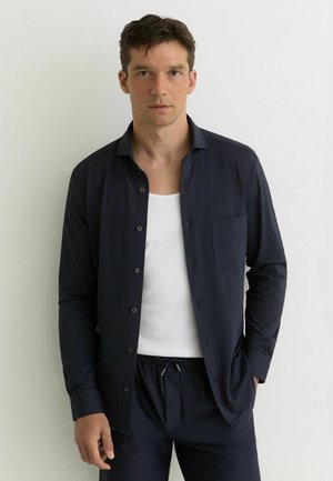 Noor noormees kannab avatud tumelilla särki valge tanktopi kohal ning sobivaid tumelillaid pükse, seisab heleda seina taustal.