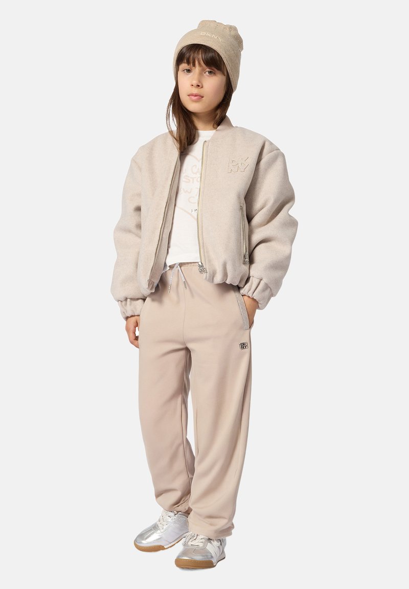 Jeune personne portant une veste polaire beige, pantalon assorti et bonnet, baskets blanches, debout avec une main dans la poche sur un fond uni.