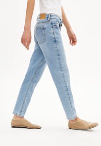 Jeans en denim bleu clair avec coupe droite, cinq poches et un patch en cuir à l'arrière. Modèle portant des chaussures à plat beige.