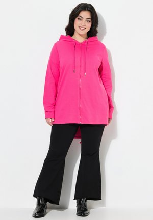 Sweat à zip rose avec cordons, manches longues et dos plus long. Associé à un pantalon noir évasé et des bottes noires. Tissu lisse.