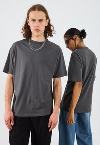 Graues, übergroßes T-Shirt aus weichem Stoff, mit rundem Hals, kurzen Ärmeln und einem minimalistischen Design. Das Model trägt silberne Kettenakzente.