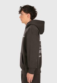 Sudadera de color marrón oscuro con bolsillo canguro en el frente, diseño tipo pullover y texto impreso en blanco en el pecho y la manga. Material de algodón suave.