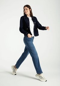 Donkere spijkerjack met twee voren zakken, wit t-shirt, blauwe jeans en lichtgekleurde sneakers. Model loopt tegen een effen achtergrond.