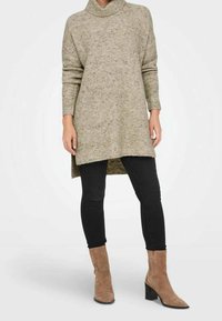 Beige gestrickte Tunika mit hohem Kragen, mit gerippter Textur, abgerundetem Saum und kombiniert mit schwarzen Skinny-Jeans und braunen Stiefeletten.