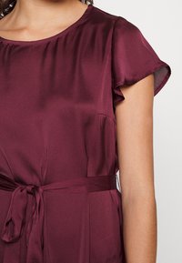 Bordeaux blouse van gladde stof met korte, gerimpelde mouwen, een ronde halslijn en een strikceintuur voor aanpassing. Heeft zijzakken.