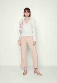 Top branco texturizado, calças largas bege, bolsa de palha bege e saltos quadrados xadrez claro. Modelo está posicionada contra um fundo neutro.