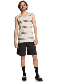Camiseta sin mangas beige con rayas horizontales verdes, pantalones cortos negros, calcetines negros y sandalias de dedo. Atuendo casual con un diseño y materiales sencillos.