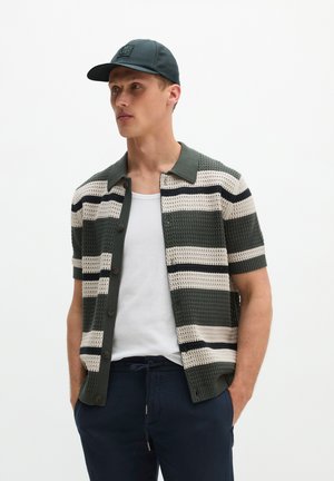 Man met een zwarte pet, kort mouw groene en witte gestreepte gebreide trui over een wit hemdje, met donkerblauwe broek, staand met handen in de zakken.
