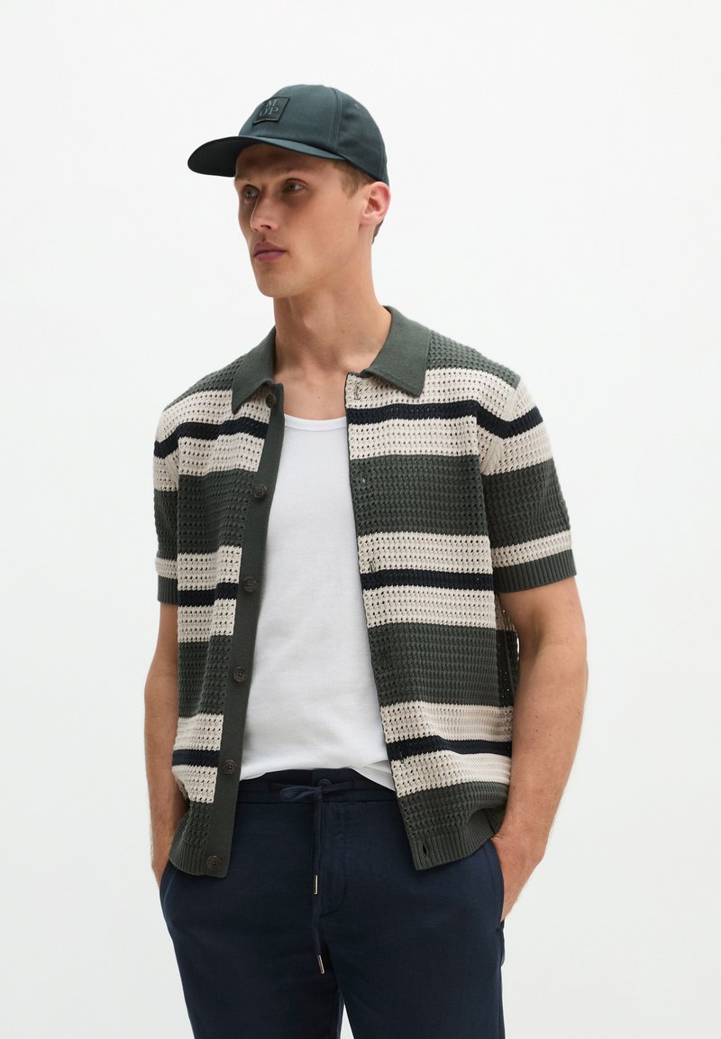 Homme portant une casquette noire, un pull rayé vert et blanc à manches courtes sur un débardeur blanc, avec un pantalon bleu foncé, debout les mains dans les poches.