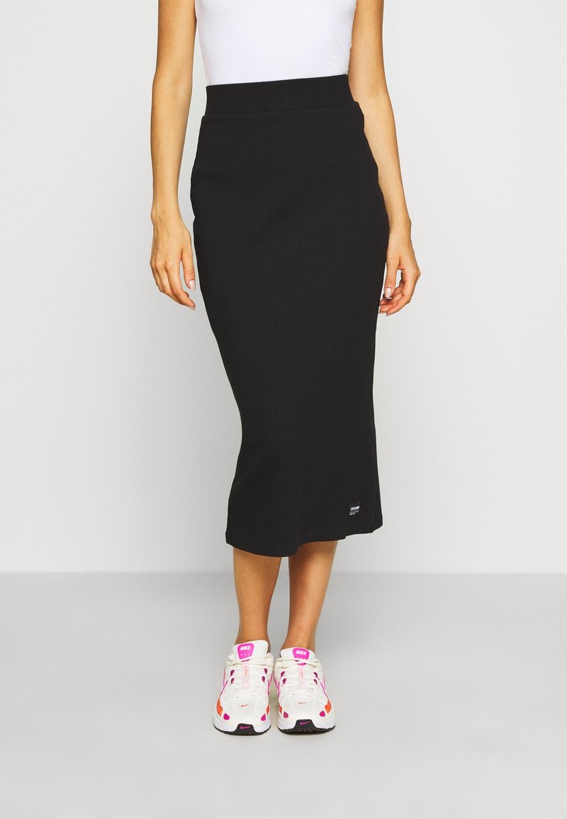 Femme portant une jupe midi noire cintrée, un top blanc sans manches et des baskets blanches avec des détails roses et orange, debout devant un fond uni.