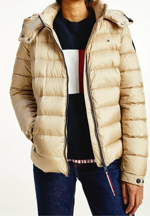 Person trägt eine beige gesteppte Steppjacke über einem marineblauen Pullover mit roten, weißen und marineblauen Streifen, kombiniert mit dunkelblauen Jeans.