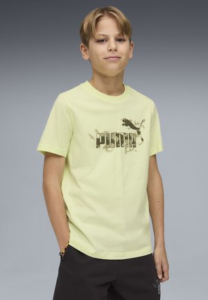 Chłopiec o krótkich blond włosach ubrany w jasną żółtą koszulkę z logo Puma i czarne spodnie, z jedną ręką w kieszeni, na tle szaro-gradientowym.