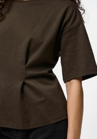 Tumši brūns īsās piedurknes T-krekls ar brīvu piegriezumu, ar sakrāto dizainu uz sāniem un apaļu kakla izgriezumu. Kokvilnas materiāls, gluda tekstūra.