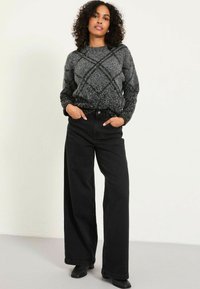 Frau mit lockigem Haar, die einen grau gemusterten Pullover, weite schwarze Hosen und schwarze Stiefel trägt, steht mit Händen in den Taschen.