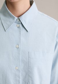 Hele sinine nööpidega särk, millel on väljakujundatud krae, väike rinnatasku ja ümar ääris. Pehme, tekstuuriga kangas nuppudega.