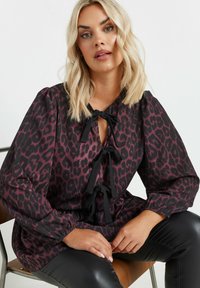 Leopardikuosisy topi tummanpunaisena ja mustana, jossa on avainreikäkaulus mustilla nauhoilla ja puffihuput, yhdistettynä kiiltäviin mustiin housuihin.