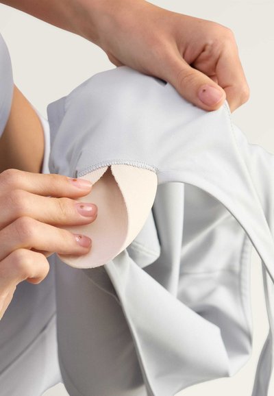 Vaaleanharmaa urheiluvaatekangas, jossa on pehmeää, nude-väristä pehmustetta, jota säädetään. Ompeleet ovat siistejä, mikä korostaa saumatonta muotoilua.