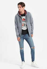 Felpa grigia con fodera a quadri rossi e lacci neri, indossata sopra una maglietta con grafica, abbinata a jeans blu strappati e sneakers bianche.