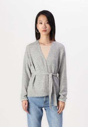 Cardigan grigio a maglia con design avvolgente, vita legata e polsini a costine con dettagli in perle. Indossato sopra una maglietta beige e jeans blu.