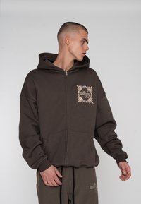 Brun zip-up hoodie med en stor frontlomme, der har et lysebrunt blomstermotiv på brystet og en hætte. Blødt, struktureret stof.