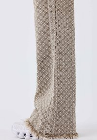 Pantalones anchos beige con superficie texturizada, con un dobladillo con flecos y un material suave y tejido. Combinados con zapatillas blancas.