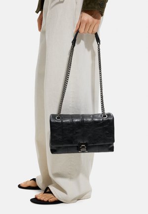 Osoba ubrana w beżowe spodnie z szerokimi nogawkami i czarne sandały, trzymająca czarną skórzaną torebkę na ramię z metalowym łańcuszkiem i zapięciem.