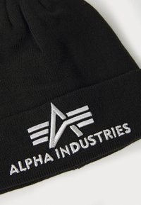 Gorro de punto negro con puño enrollado. Presenta el texto bordado en blanco "ALPHA INDUSTRIES" y un logo con rayas y ángulos estilizados.