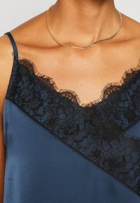 Gros plan d'une personne portant un caraco en satin bleu marine avec bordure en dentelle noire et un collier chaîne en or sur un cou et des épaules nus.