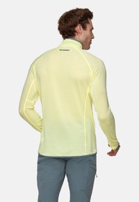 Mann, der ein hellgelbes Langarmoberteil mit "Mammut"-Logo auf dem oberen Rücken und grauen Hosen trägt, stehend mit dem Rücken zur Kamera vor einem weißen Hintergrund.