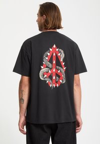 Czarny bawełniany t-shirt z odważnym, czerwono-białym wzorem graficznym przedstawiającym węża owiniętego wokół abstrakcyjnego kształtu.
