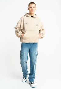 Beżowa bluza z kapturem z przednią kieszenią i logo, w połączeniu z jasnoniebieskimi spodniami cargo z bocznymi kieszeniami i luźnym krojem, uzupełniona o trampki.