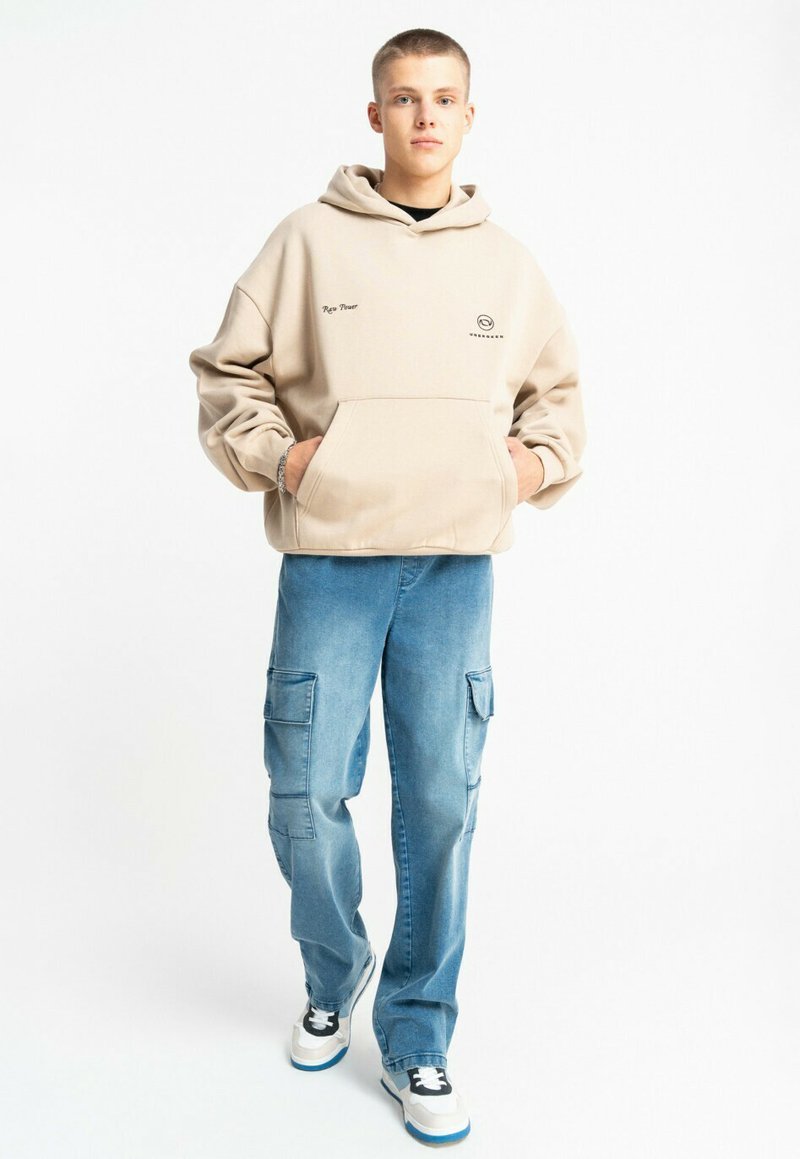 Beżowa bluza z kapturem z przednią kieszenią i logo, w połączeniu z jasnoniebieskimi spodniami cargo z bocznymi kieszeniami i luźnym krojem, uzupełniona o trampki.